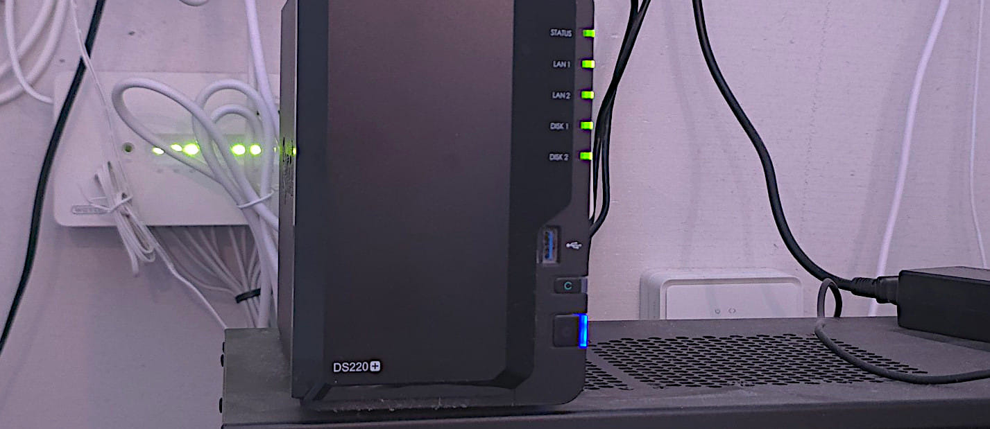 A Synology DS220+ NAS on a shelf, status and disk LEDs lit green, cabled to a network switch behind it — the machine the mirror cron runs on